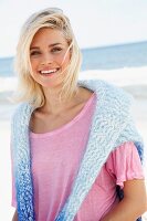 A blonde woman oon a beach wearing a pink t-shirt and a jumper over her shoulders
