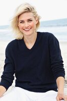 A blonde woman on a beach wearing a dark woollen jumper and white trousers