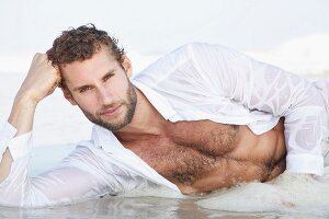 Junger Mann in nassem, weissen Hemd liegt am Strand