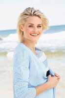 A blonde woman on a beach wearing a striped shirt and a light blue cardigan