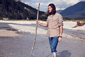 A young man with a beard and long hair walking along a gravelly river bed