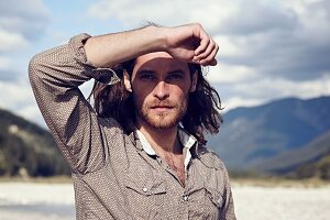 A young man with a beard and long hair walking along a gravelly river bed