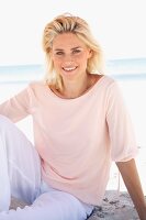 Lächelnde blonde Frau in pastellfarbenem Shirt und Hose sitzt am Strand