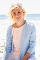 Lächelnde blonde Frau in pastellfarbenem Shirt, Jacke und Hose sitzt am Strand