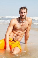 A muscular young man wearing yellow Bermuda shorts sitting in the shallows on a beach