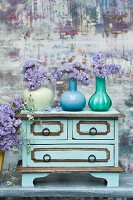 Dried, purple flowers in three colourful, glass vases on top of vintage chest of drawers against painted background