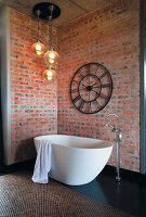 Freistehende Badewanne vor Backsteinwand mit großer Wanduhr und Kugelleuchten