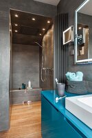 A petrol-coloured washstand with a grey-tiled shower in the background with built-in spot light in the ceiling