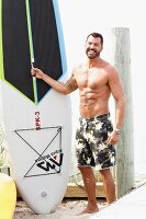 A dark-haired man wearing bathing shorts holding a surfboard