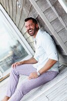 A dark-haired man with a beard wearing a white shirt and checked trousers sitting on the floor laughing