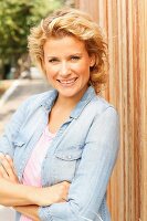 A blonde woman wearing a denim shirt leaning on a fence