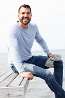 A man wearing a grey knitted hat, a light-blue jumper and jeans by the sea