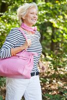 A blonde woman in the countryside wearing a striped top and white trousers holding a pink bag