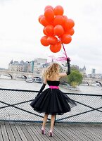 A young woman on a bridge wearing a black petticoat dress with a pink belt and holding a bunch of red balloons