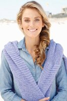 A young blonde woman on a beach wearing a denim shirt with a jumper over her shoulders