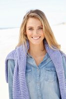 A young blonde woman on a beach wearing a denim shirt with a jumper over her shoulders