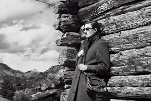 A short-haired woman wearing sunglasses, a knee-length coat and a turtleneck jumper in front of a wooden hut (black-and-white shot)