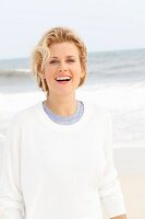 A blonde woman on a beach wearing a blue top and a white round-neck jumper