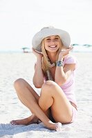 A young blonde woman sitting on a beach wearing a summer hat, scarf and pink T-shirt