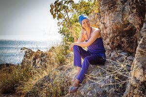 Frau in violettem Ensemble und Turban sitzt an Felsen am Meer