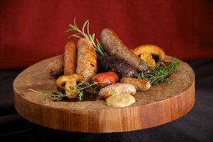 A wooden platter with various cooked sausages, herbs, mushrooms and mustard