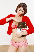 Junge Frau in Unterwäsche und Strickjäckchen nascht aus großem Glas Nutella