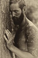 A topless man with a large tattoo leaning against a tree