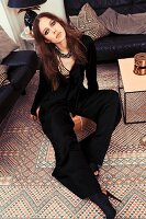 A young woman wearing a wide black jumpsuit sitting on a leather stool