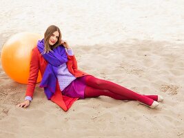 Frau mit Winterkleidung in Rot- und Lilatönen sitzt im Sand