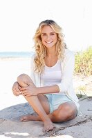 A young blonde woman on a beach wearing a white top, jacket and shorts