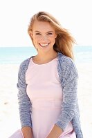A young blonde woman on a beach wearing a pastel pink dress and a cardigan