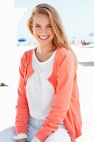 Blonde Frau in weißem Pulli und lachsfarbener Strickjacke am Strand