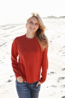 A young woman on a beach wearing a rust-red jumper and jeans