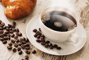 A steaming cup of coffee with coffee beans and a croissant on a light wooden surface