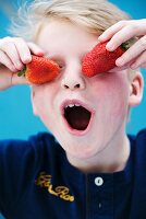 A boy holding two strawberries in front of his eyes