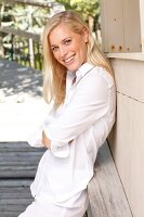 A young blonde woman leaning on a wooden facade wearing a white blouse