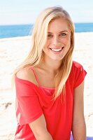 A young blonde woman sitting on a beach wearing a red top