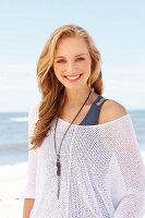 A young blonde woman on a beach wearing a tank top and a net jumper