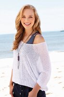 A young blonde woman on a beach wearing a tank top and a net jumper
