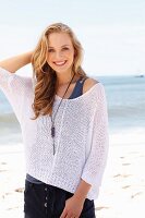 A young blonde woman on a beach wearing a tank top and a net jumper