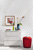 A red upholstered stool next to a wallpapered chest of drawers in front front of wallpaper with the same grey tile pattern