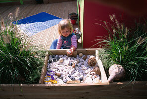 CHILDRENS DECK Garden: Lucy FEELING THE SHELLS, FIR CONES AND STONES IN A RAISED WOODEN BED