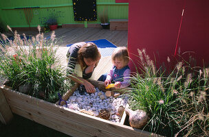 CHILDRENS DECK Garden: Lucy AND Clare FEELING THE SHELLS, FIR CONES AND STONES IN A RAISED WOODEN BED: Designer: Clare MATTHEWS