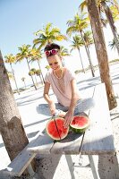 A brunette woman wearing a cream T-shirt and a pair of light grey trousers slicing a watermelon on the beach