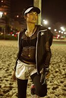 Dunkelhaarige Frau mit Kappe, Sport-BH, schwarzem Netzshirt, Sportjacke, silbernen Shorts und Leggings am Strand