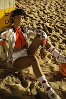 A dark-haired woman sitting on a beach and wearing a cropped top, silver sports jacket and shorts