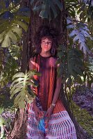 A brunette woman wearing a pleated top, a patterned pleated skirt and holding a clutch with tropical print