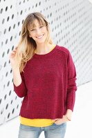 Blonde Frau in gelbem T-Shirt, rotem Strickpullover und Jeans vor Wand mit Löcher