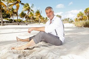A man with grey hair wearing a white shirt and grey trousers sitting on the beach