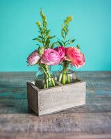 Glasfläschchen mit Ranunkelblüten in Holzkistchen vor türkisfarbenem Hintergrund